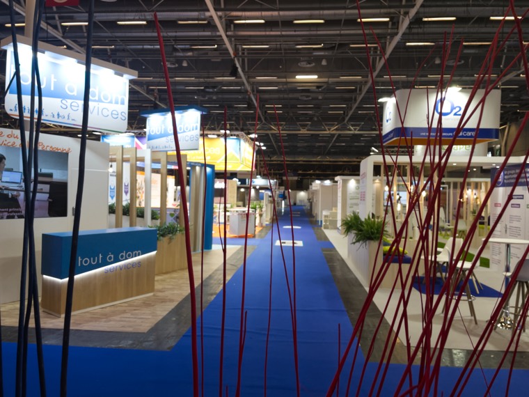 Allée d’un salon professionnel bordée de stands, moquette bleue